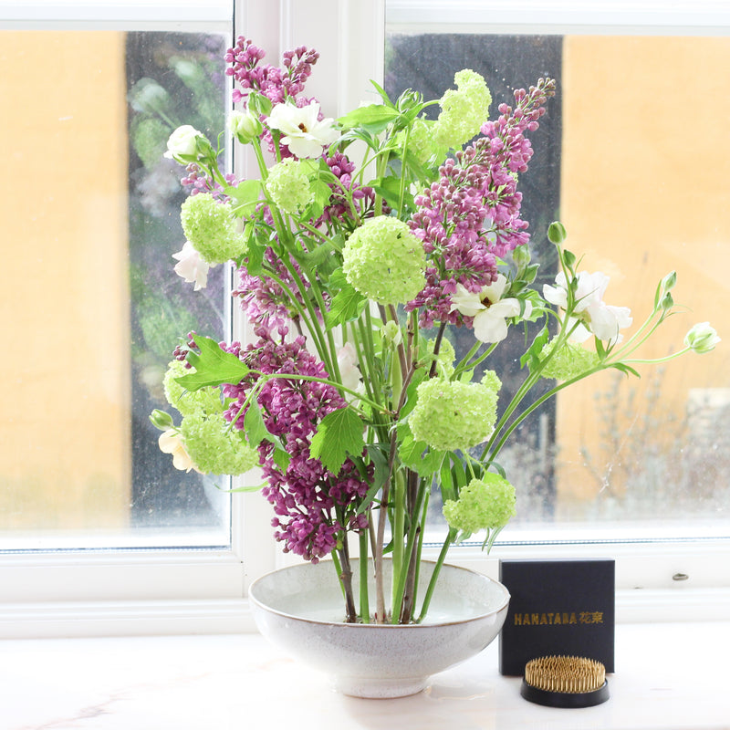 A vibrant display of lush green Viburnum and delicate purple lilacs arranged in a textured grey bowl, by a window overlooking a yellow building, with a small black HANATABA kenzan box and a golden kenzan nearby, creating a cozy and inviting indoor garden scene