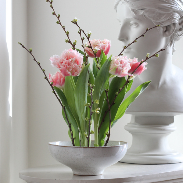 Élégant arrangement floral composé de tulipes roses frangées et de délicates branches bourgeonnantes dans un bol moucheté, juxtaposé à un buste blanc classique.