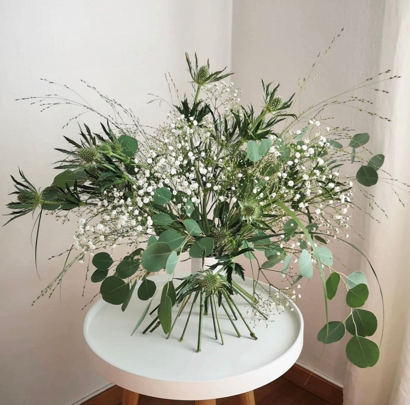 Arrangement ikebana élégant avec un assortiment de verdure et de fleurs blanches fixé par un Hanataba Bouquet Twister en argent nacré sur une table blanche.