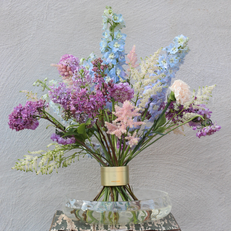 Élégant arrangement floral Ikebana composé d'une variété de fleurs aux couleurs pastel, dont des lilas et des delphiniums, fixé par un chic support de tiges en spirale.