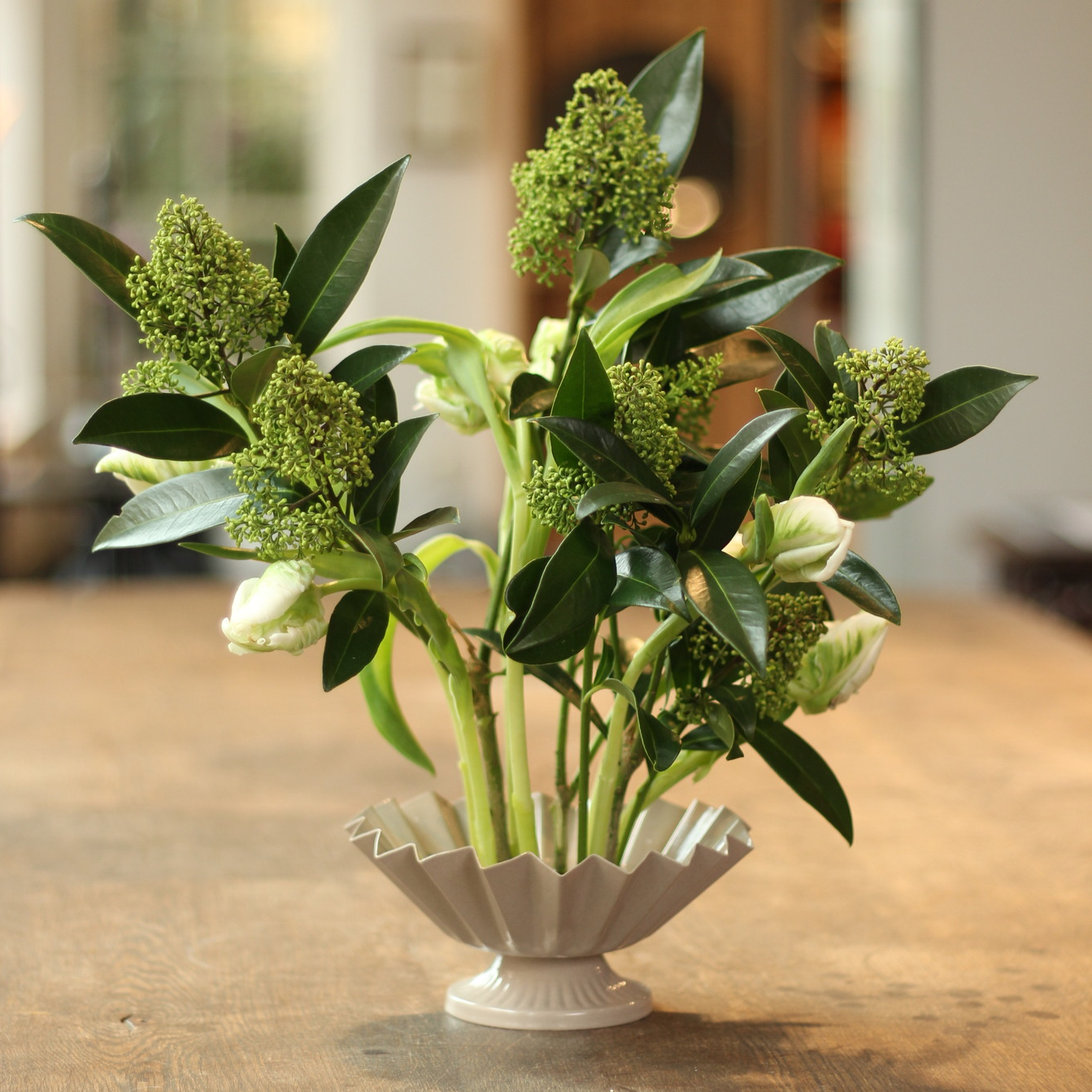 Elegant floral arrangement featuring green budding flowers and lush leaves in a classic white fluted vase anchored on a kenzan ring, presented on a wooden surface with a softly blurred background.