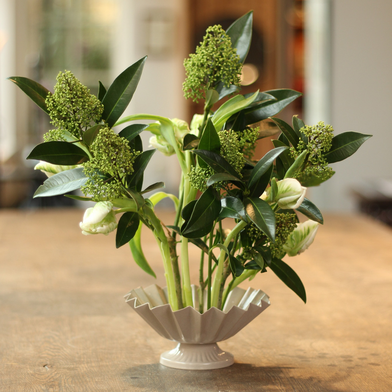 Arrangement floral élégant composé de fleurs vertes en bouton et de feuilles luxuriantes dans un vase cannelé blanc classique ancré sur un anneau de kenzan, présenté sur une surface en bois avec un arrière-plan légèrement flou.