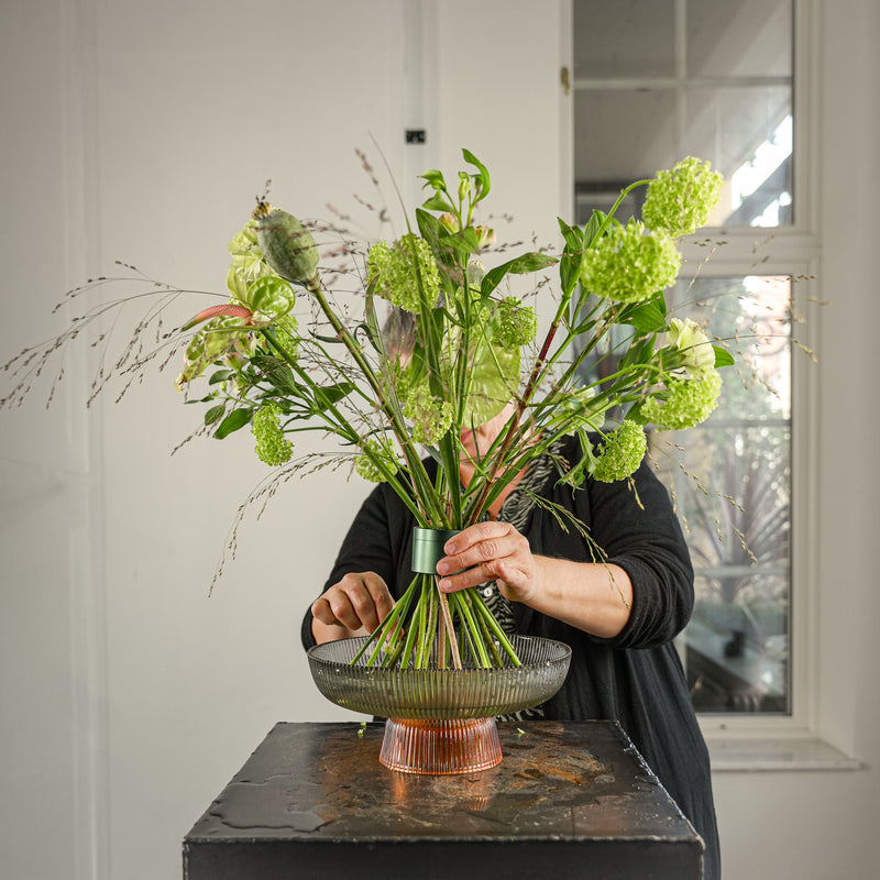 Un fleuriste crée un arrangement floral professionnel avec des fleurs coupées de viburnum dans un Hanataba Ivy Green, présentées dans un plateau de verre rond rayé.