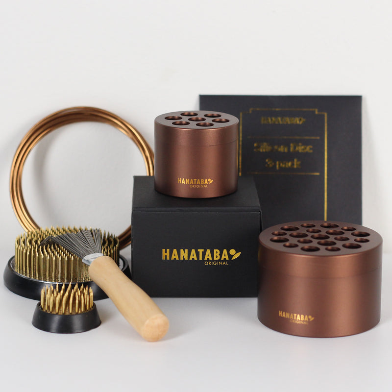 Un ensemble d'outils de composition florale Hanataba Original, comprenant deux porte-fleurs marron, un grand kenzan (70 mm), un petit kenzan (34 mm) et une brosse à râteau kenzan avec un manche en bois. Les articles sont présentés dans une boîte noire avec le logo Hanataba en lettres dorées.