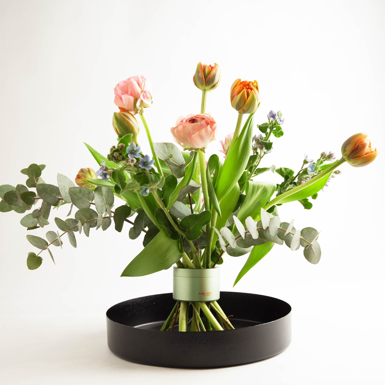 Magnifique bouquet de tulipes roses et orange et de feuilles d'eucalyptus disposées dans un Twister de fleurs vertes Hanataba Matcha, placé dans un vase rond en métal noir entouré d'un blanc éclatant.