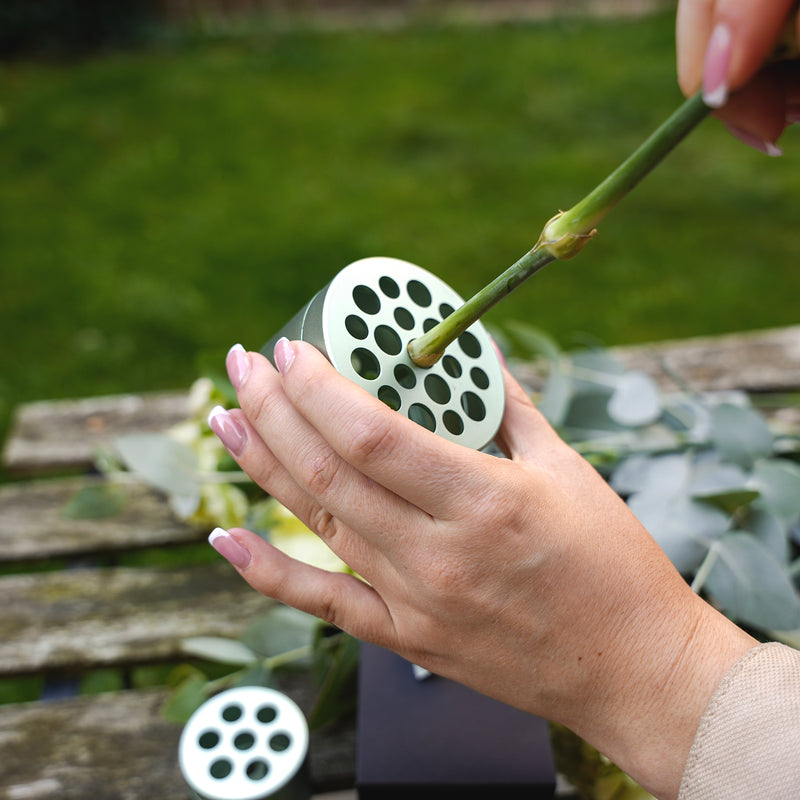 Une personne avec du vernis à ongles rose arrangeant des eucalyptus et des fleurs en insérant une tige dans un outil de torsion de bouquet Hanataba Matcha Green au-dessus d'une table en bois dans le jardin.