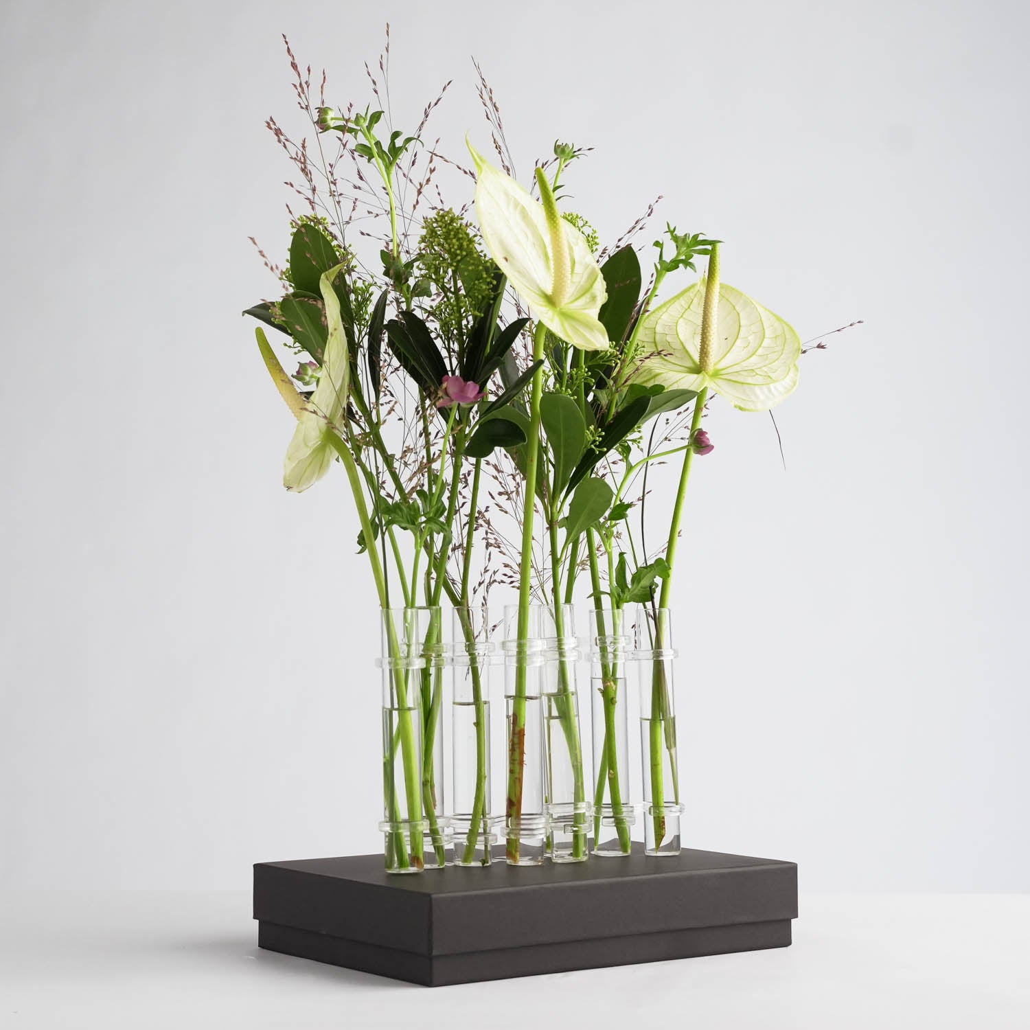 A minimalist floral arrangement featuring tall, slender green and white flowers, including anthuriums and greenery in glass flower flute vases on a rectangular black packaging box, set against a plain white background.