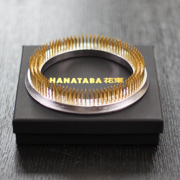 Une bague Hanataba kenzan dorée avec une base en argent, présentée dans un coffret noir avec texte en or, mettant en valeur d'élégants outils d'art floral japonais.