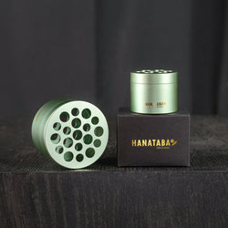 Deux torsades de bouquet Hanataba dans le design Matcha Green. Les produits sont placés dans leur emballage d'origine noir avec Hanataba Original écrit en or. L'exposition se fait dans une pièce sombre sur une table en bois noir.