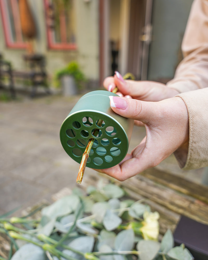 Une personne tenant une grenouille florale Hanataba Ivy Green avec une tige de fleur insérée dans l'un de ses trous. Des feuilles d'eucalyptus et d'autres plantes vertes sont éparpillées sur la surface. L'arrière-plan montre un environnement extérieur.