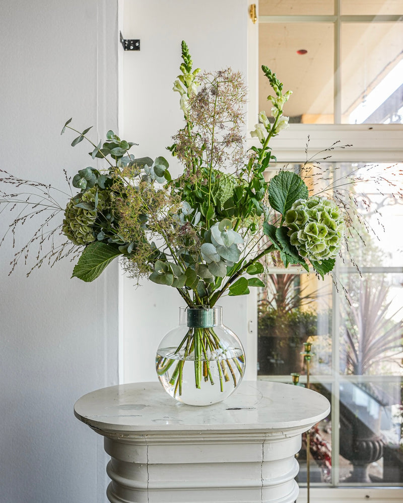 Un vase en verre transparent contenant une composition de fleurs vertes et blanches, dont des hortensias et des eucalyptus, est posé sur une petite table ronde blanche. Le bouquet Hanataba Ivy Green est mis en valeur par la lumière du soleil qui traverse une grande fenêtre à l'arrière-plan.