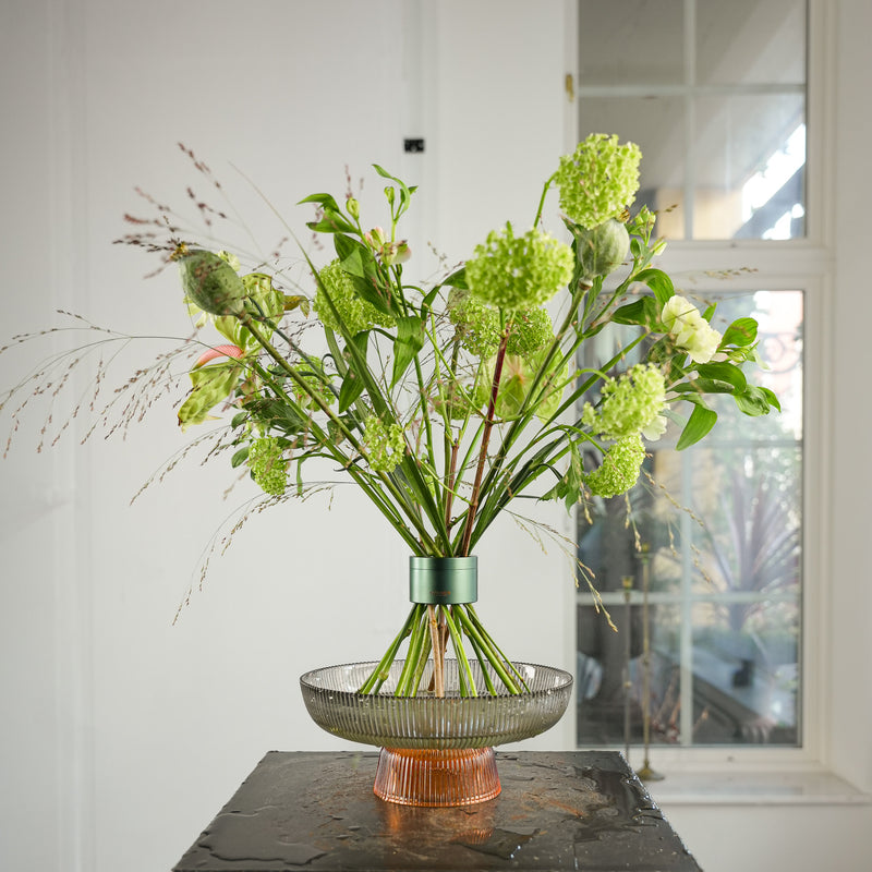 Superbe bouquet de fleurs de graminées et de viburnum arrangé dans un outil Hanataba Ivy Green, présenté dans un plateau en verre avec de l'eau sur une table noire près d'une fenêtre dans une pièce blanche et lumineuse.