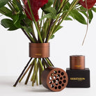 Muddy brown ikebana-inspired floral arrangement tool showcasing the cylindrical Hanataba, one with visible stem-holding holes, placed next to greenery. Another set rests on a black packaging box labeled with HANATABA logo in golden letters, creating an elegant bouquet display against a white background.