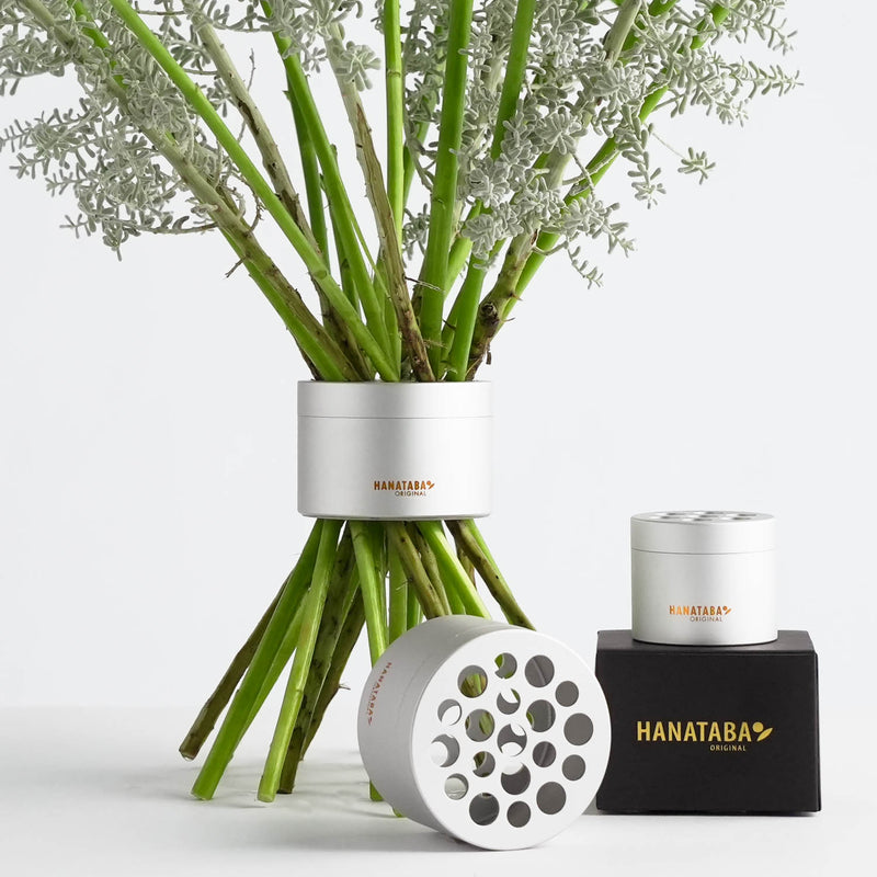 A white flower arrangement showcasing green stems, paired with two more holders on a black box labeled HANATABA ORIGINAL. The floral design features circular cutouts, ideal for arranging stems and twisting the flowers into a Dutch spiral bouquet.