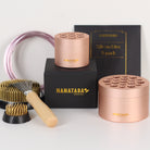 A set of Hanataba Original flower arrangement tools, featuring two Hanataba Pink Champagne floral holders, a larger kenzan (70mm), a smaller kenzan (34mm) and a kenzan rake brush with a wooden handle. The items are arranged on a black box with golden Hanataba logo lettering.