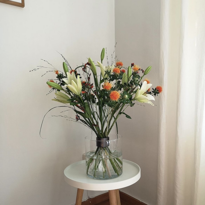 Arrangement floral minimaliste de style Ikebana composé de lys blancs et de Carthamus orange dans un vase transparent avec un support de tige en spirale Hanataba sur une table blanche.