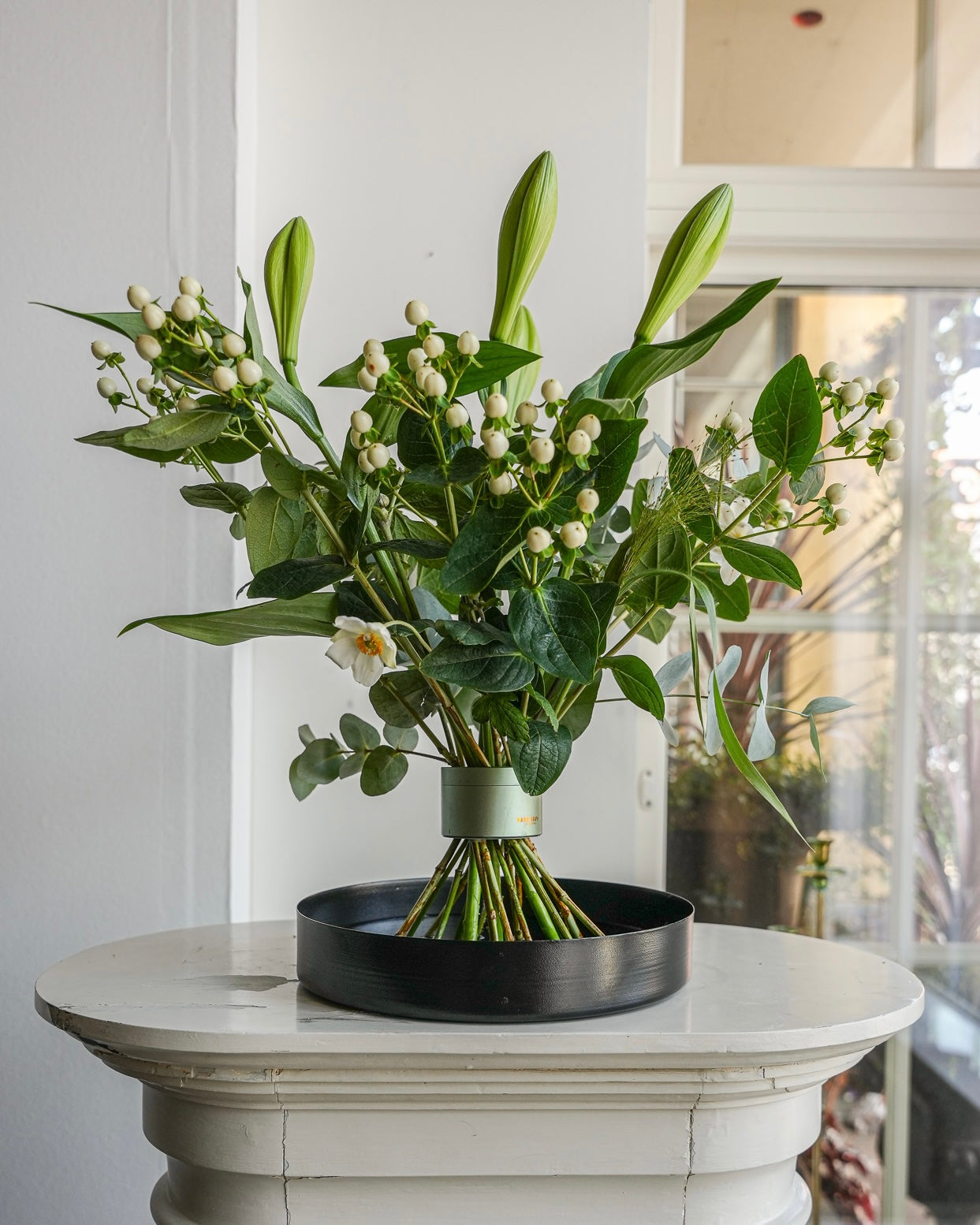 Stunning flower arrangement with lilies displayed on a black floral tray atop a white pedestal in a beautiful white room with paneled windows.