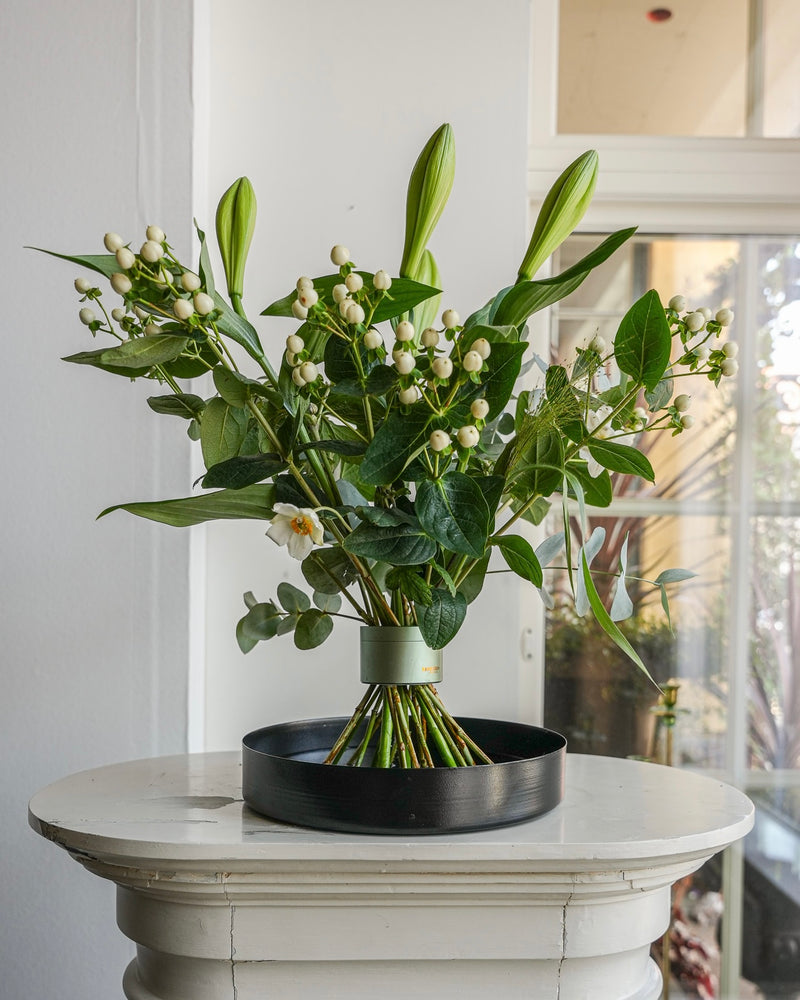 Superbe composition florale de lys présentée sur un plateau floral noir surmontant un piédestal blanc dans une belle pièce blanche aux fenêtres lambrissées.