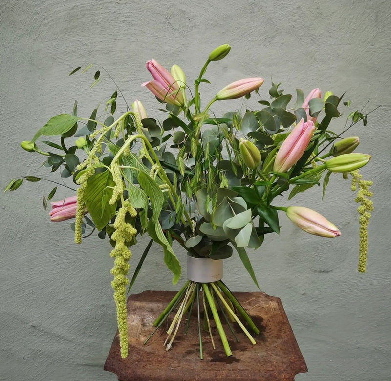 Un arrangement floral luxuriant composé de lys roses et d'accents verts en cascade, le tout maintenu par un Hanataba Bouquet Twister en argent nacré, sur un mur gris texturé.