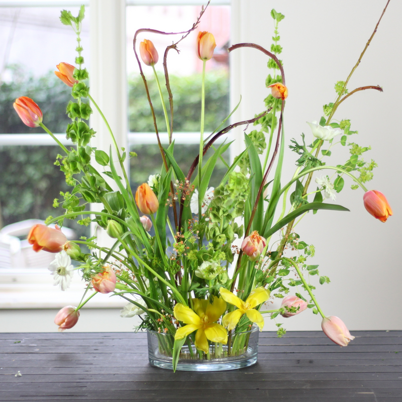 Un arrangement vibrant de tulipes et de verdure mixte se tenant debout dans un anneau de kenzan de 200 mm, placé sur une table en bois sombre près d'une fenêtre.