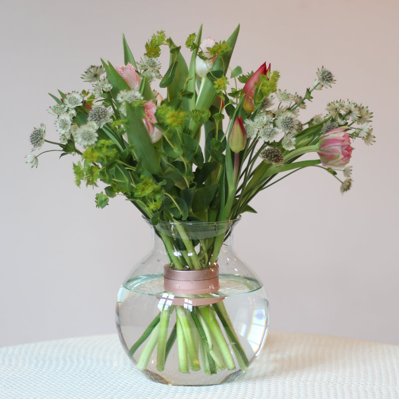 Un bouquet frais de tulipes et de verdure assortie, maintenu par une torsade de bouquet Hanataba rose nacré, est placé dans un vase rond transparent, offrant un mélange d'élégance et de charme printanier.