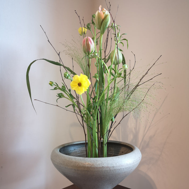  Une composition florale sereine avec une jonquille jaune et des tulipes rose tendre, le tout fixé par un anneau Kenzan de 70 mm dans une coupe en pierre.