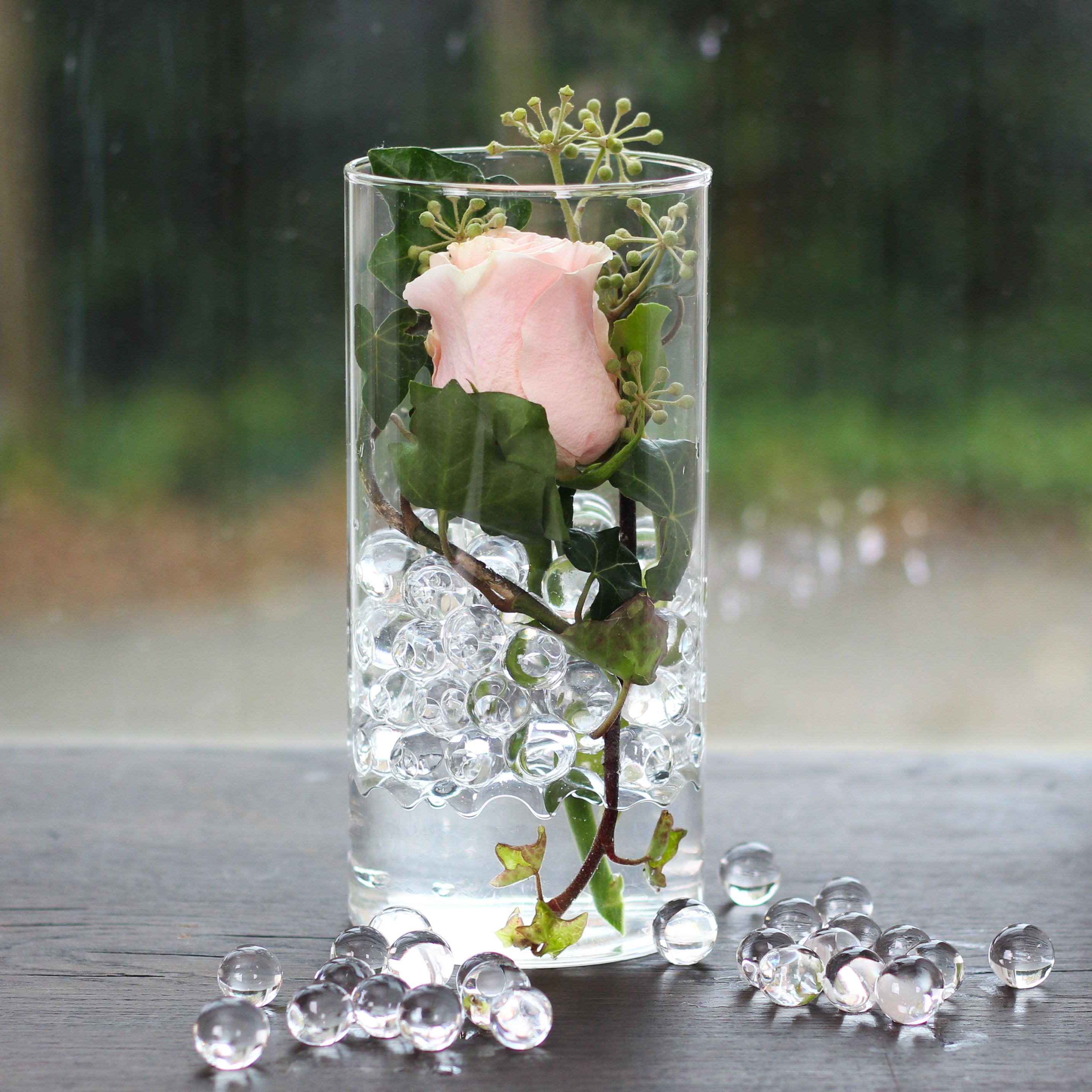 A single pale pink rose with ivy, poised in a clear vase filled with water and transparent Water hydrogel beads.