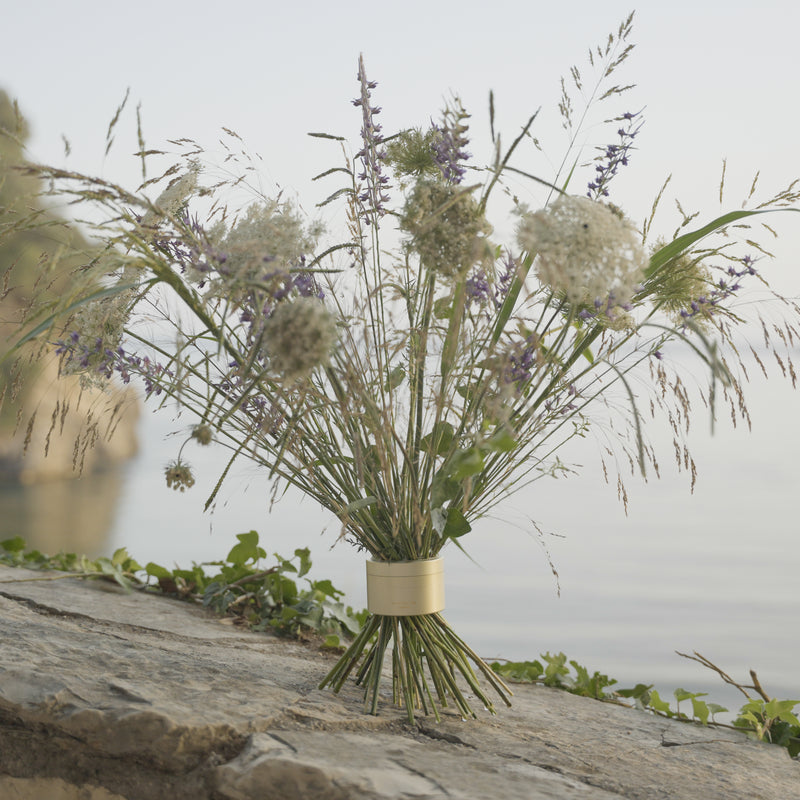 Des fleurs sauvages et des herbes dans un Hanataba Bouquet Twister Champagne Gold, posé sur un rocher avec de l'eau sereine à l'arrière-plan, qui évoque la tranquillité naturelle.